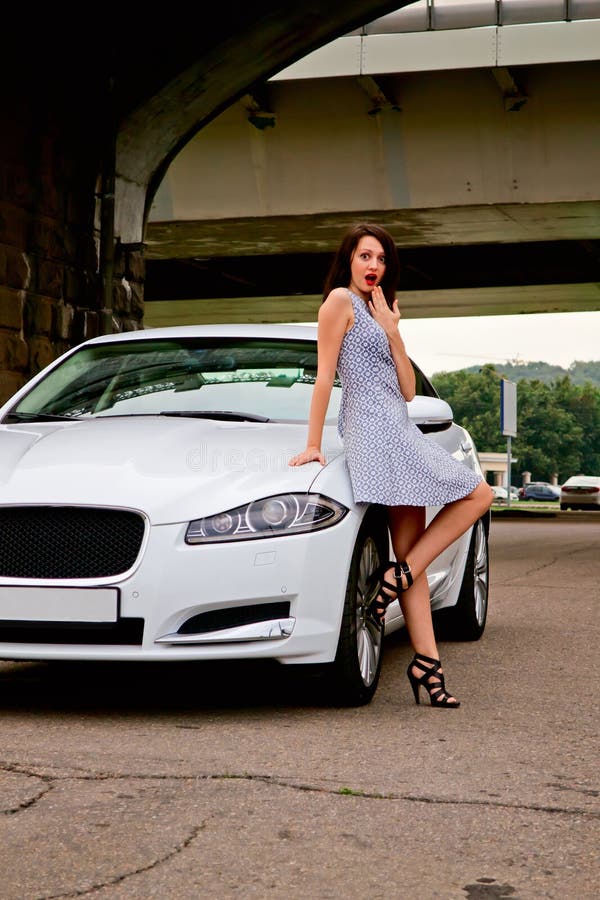 Cute brunette and luxury car. Cute brunette with red lips on high heels is posing with luxury white car (Jaguar XF) outdoor. Showing emotional oops / wow pinup royalty free stock image