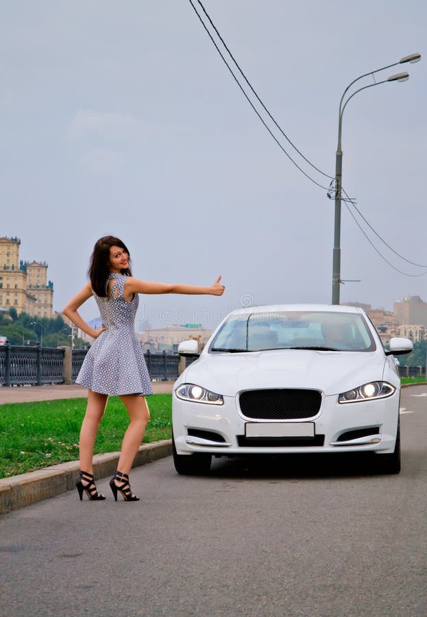 Cute brunette and luxury car. Cute brunette with red lips on high heels is stopping luxury white car (Jaguar XF) outdoor. Hitch-hiking stock image