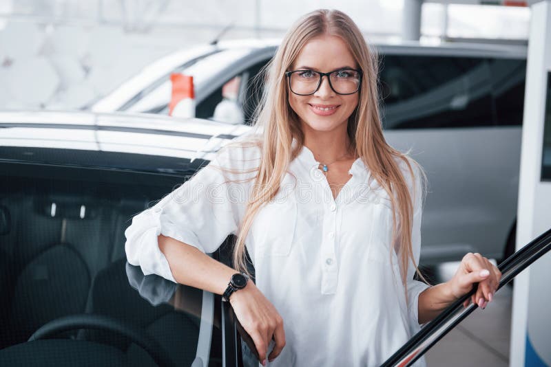 It`s her birthday gift. Cute girl in eyewear stands near the car in auto saloon. Probably her next purchase.  stock photography